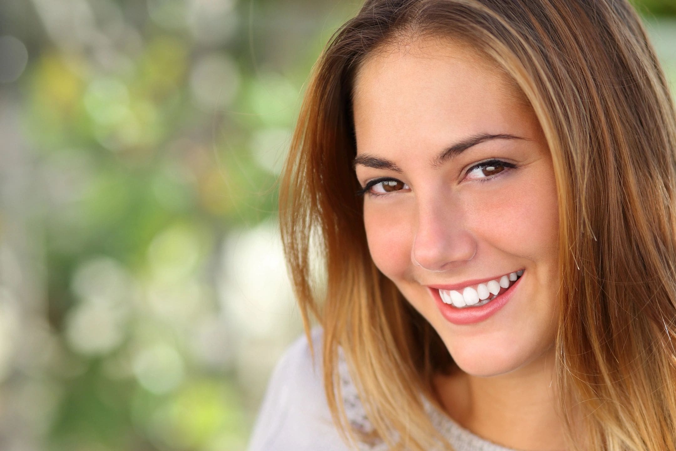 A young woman with long, light brown hair smiles at the camera with a blurred green background.
